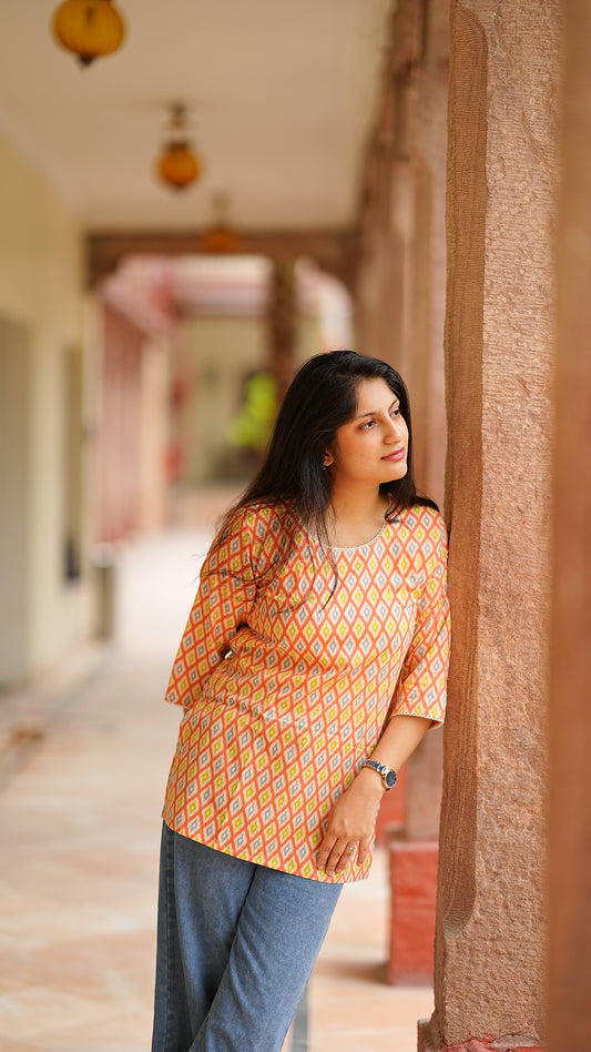 Woman in an orange patterned top standing in a sunlit corridor with architectural elements.